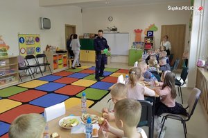 Sala przedszkolna z dziećmi przy stolikach, widoczny umundurowany policjant