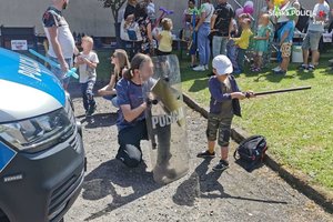 Zdjęcie przedstawiające rozproszoną grupę dzieci na tle zieleni, na pierwszym planie przód radiowozu oraz meżczyzna klęczący przy dziecku z tarczą policyjną i pałką