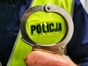 Zbliżenie kajdanek trzymanych w ręku policjanta na tle jego żółtej kamizelki odblaskowej