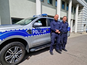 Dwóch umundurowanych policjantów na tle oznakowanego radiowozu