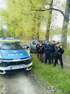 Grupa mężczyzn w mundurach, w tym policjant nad brzegiem rzeki, na pierwszym planie terenowy radiowóz