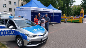 Policjant z mężczyzną i dwójka chłopców na tle namiotów wystawienniczych, na pierwszym planie radiowóz