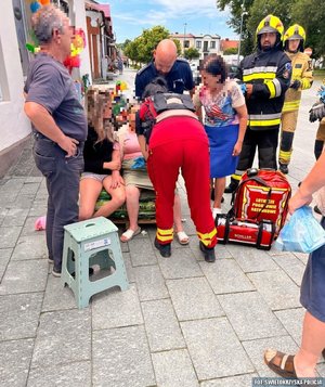 Rynek miasteczka, grupa ludzi zgromadzona wokół siedzącej na krześle starszej kobiety, widoczni strażacy, ratownicy medyczni a także policjant