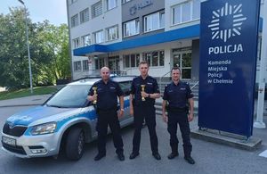 Trzech umundurowanych policjantów na tle komendy Policji i oznakowanego radiowozu