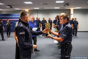 Sala konferencyjna policjant wręczający puchar drugiemu policjantowi, w tle inni policjanci