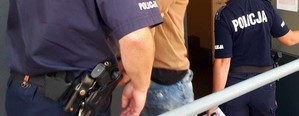 Zbliżenie od tyłu dwóch policjantów oraz mężczyzny ze skutymi z tyłu rękami