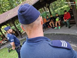 Dwaj policjanci widziani od tyłu a pod wiatą letniskową grupka młodzieży