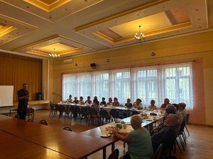 Sala konferencyjna, przy stolikach seniorzy, centralnie policjant prowadzący spotkanie