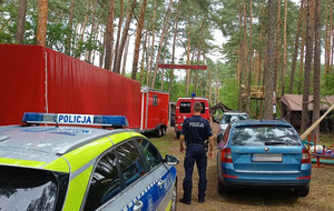 Obóz harcerski w lesie na pierwszym planie widoczny radiowóz oraz policjant dalej wozy straży pożarnej