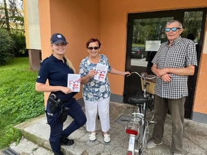 Dwójka seniorów z rowerem przed klatką schodową obok nich uśmiechnięta policjantka