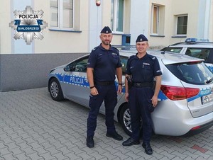 Dwoch uśmiechniętych policjantów obok radiowozu zaparkowanego przy komisariacie