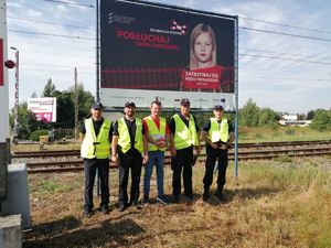 Pięciu mężczyzn w odblaskowych kamizelkach na tle torów kolejowych pod bilbordem z wizerunkiem dziewczynki i napisem &quot;Posłuchaj głosu rozsądku, zatrzymaj się przed przejazdem tatusiu&quot;