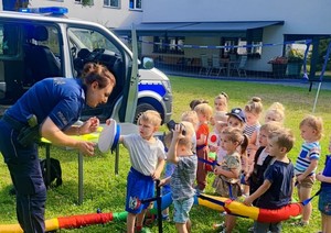 Policjantka pochylająca się do grupki dzieci na tle radiowozu