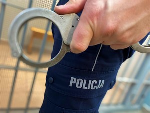 Zbliżenie kajdanek trzymanych w dłoni umundurowanego policjanta na wysokości nogi