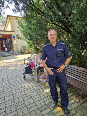 Umundurowany policjant na tle zieleni i dalej budynku