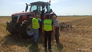 Umundurowany policjant w żółtej kamizelce z napisem Policja z dwoma mężczyznami na polu na tle ciągnika rolniczego