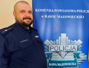 Zdjęcie umundurowanego policjanta na tle baneru Komendy Powiatowej policji w Rawie Mazowieckiej