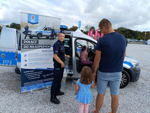 policjantka obok radiowozu i baneru policji, przed nią mężczyzna z dzieckiem