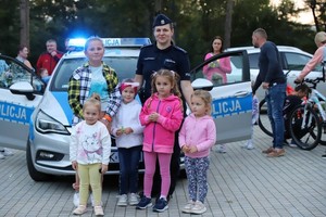 Pozująca do zdjęcia policjantka z grupką dzieci na tle radiowozu