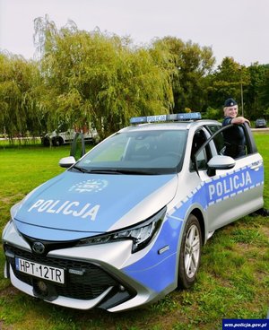 Uśmiechnięta policjantka bok radiowozu na tle zieleni miejskiej