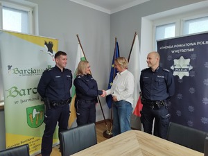 Trójka policjantów oraz kobieta w białej bluzce ściskająca dłoń na tle granatowych flag