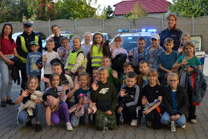 Grupka dzieci pozująca na zewnątrz do zdjęcia, widoczny policjant, w tle radiowóz