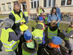 Policjant z dziećmi w odblaskowych kamizelkach na tle budynku