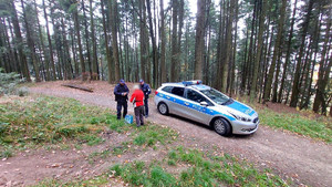 W scenerii leśnej widziane z dala: radiowóz, dwóch policjantów i starsza kobieta