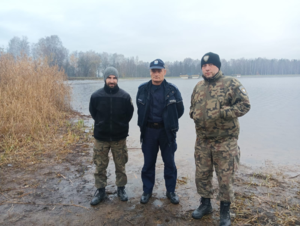 Policjant oraz dwóch mężczyzn ubranych w stroje typu panterka nad brzegiem zbiornika wodnego