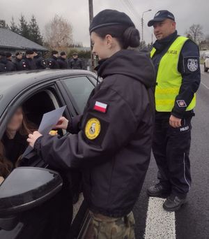 Dziewczyna ubrana w &quot;kurtkę mundurową&quot; dająca przez okno w drzwiach samochodu kartkę kierującemu, za nią policjant