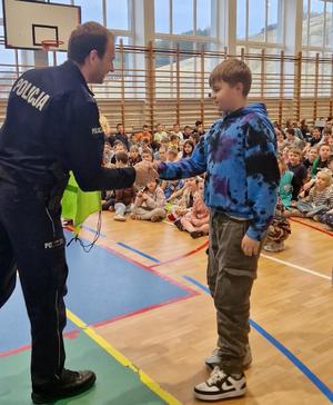 Policjant ściskający rękę chłopca na sali gimnastycznej wypełnionej dziećmi i młodzieżą