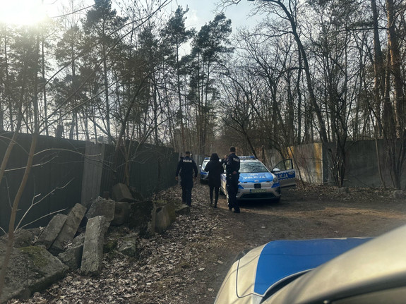 Droga miedzy dwoma betonowymi plotami w zaroślach w oddali widoczny radiowóz, dwóch policjantów i kobietę