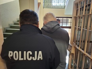 Widziani od tyłu policjant oraz mężczyzna po cywilnemu stojący w korytarzu bok krat aresztu