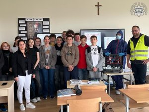 Grupa młodzieży pozująca do zdjęcia w sali szkolnej, wśród nich osoba w kombinezonie jednorazowym z walizką kryminalistyczną oraz mężczyzna w kamizelce odblaskowej
