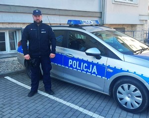 Policjant pozujący do zdjęcia na tle radiowozu i ściany budynku