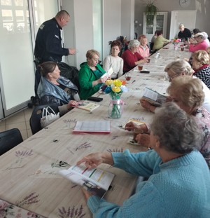 Seniorzy siedzący przy stole w świetlicy, a przy nich pochylny dzielnicowy