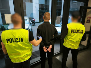 Widziani z tylu dwaj policjanci w kamizelkach odblaskowych wyprowadzający z budynku mężczyznę ze skutymi rękami