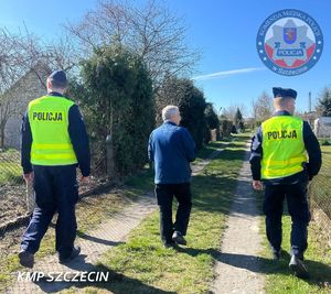Dwóch policjantów oraz mężczyzna w kurtce dżinsowej idący aleją ogródków działkowych