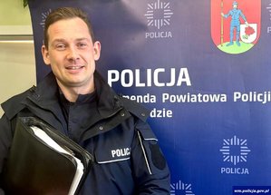 Uśmiechnięty policjant z aktówką na tle banneru swojej jednostki