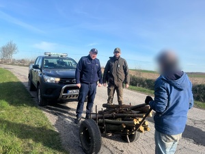 Polna droga, w tle samochód terenowy a przed nim policjant, strażnik leśny i mężczyzna z wózkiem ręcznym, na którym leżą kłody drzewa
