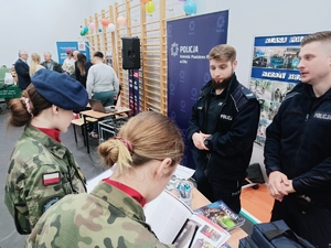 Stoisko promocyjne w sali gimnastycznej obsługiwane przez dwóch policjantów wkoło widoczna młodzież, w tym w mundurach