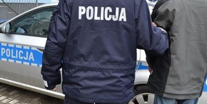 Plecy policjanta przytrzymującego mężczyznę ze skutymi rękami na tle radiowozu