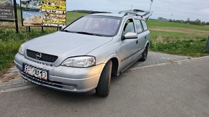 Srebrny samochód osobowy typu kombi z podniesioną pokrywa bagażnika stający na skraju posesji.