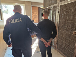 Policjant przytrzymujący mężczyznę ze skutymi z tyłu rękami obok krat aresztu