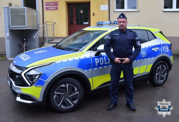 Policjant pozujący do zdjęcia na tle radiowozu i budynku komendy