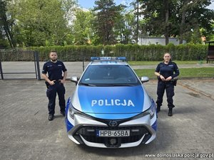Policjantka i policjant po obu stronach radiowozu stającego na placu, w tle zieleń