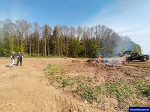 Ognisk na otwartej przestrzeni, w tle las, ładowarka oraz policjant i mężczyzna