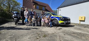 Pozujący do zdjęcia: dwójka policjantów z grupką dzieci i opiekunami na tle radiowozu