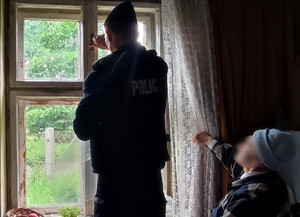 Policjant wewnątrz mieszkania przy oknie z wybitymi szybami, obok starsza kobieta