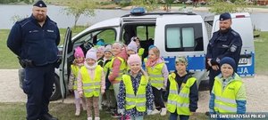 Grupa przedszkolaków w żółtych odblaskowych kamizelkach przed radiowozem, po obu stronach dwóch policjantów